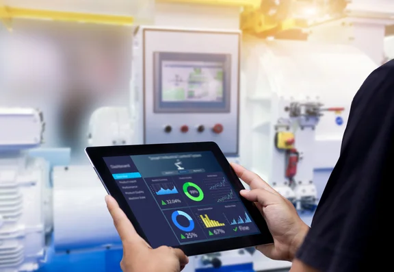 Hände, die ein Tablet vor einer verschwommener Automatisierungsmaschine halten.