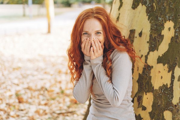 Amüsierte junge Frau kichert in die Kamera mit den Händen vors Gesicht, während sie sich in einem Park an einen Baumstamm lehnt