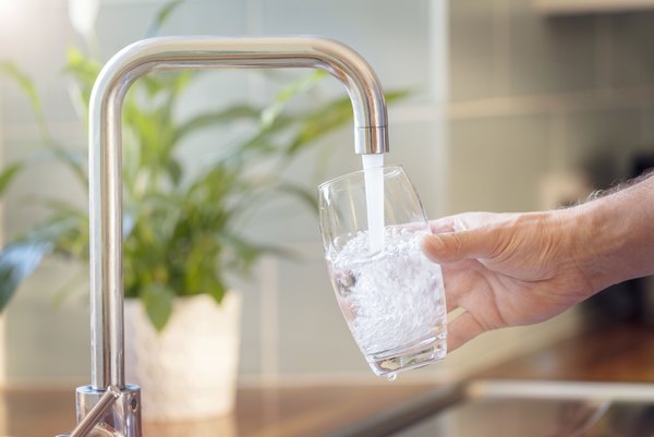 Eine Hand hält ein Glas unter den Wasserhahn in der Küche und füllt es mit Trinkwasser auf.