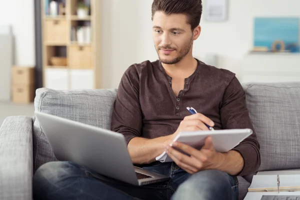 Ein junger Mann sitzt mit Laptop und Unterlagen auf dem Sofa in seiner Wohnung.