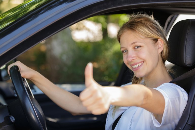 Eine lächelnde junge Frau sitzt im Auto und zeigt den Daumen nach oben.